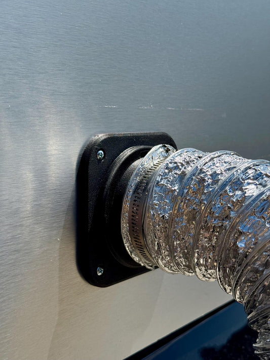 May include: A black plastic vent cover is mounted on a brushed metal surface. A flexible, corrugated silver duct is attached to the vent cover with a metal clamp. The duct is made of a shiny, metallic material. The vent cover is secured to the surface with black screws.