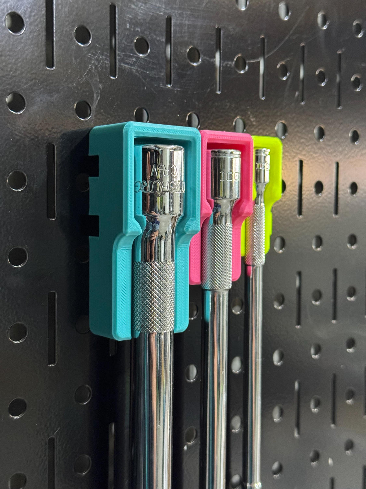 Side-angled view of three extension heads secured in teal, pink and yellow holders on black pegboard.