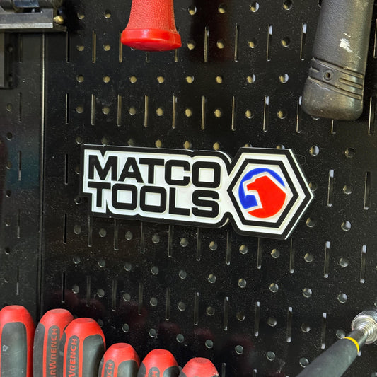 May include: A black pegboard with tools and a white and black Matco Tools logo. The logo features the text "MATCO TOOLS" and a red, blue, and white emblem. Red-handled tools are visible at the bottom.