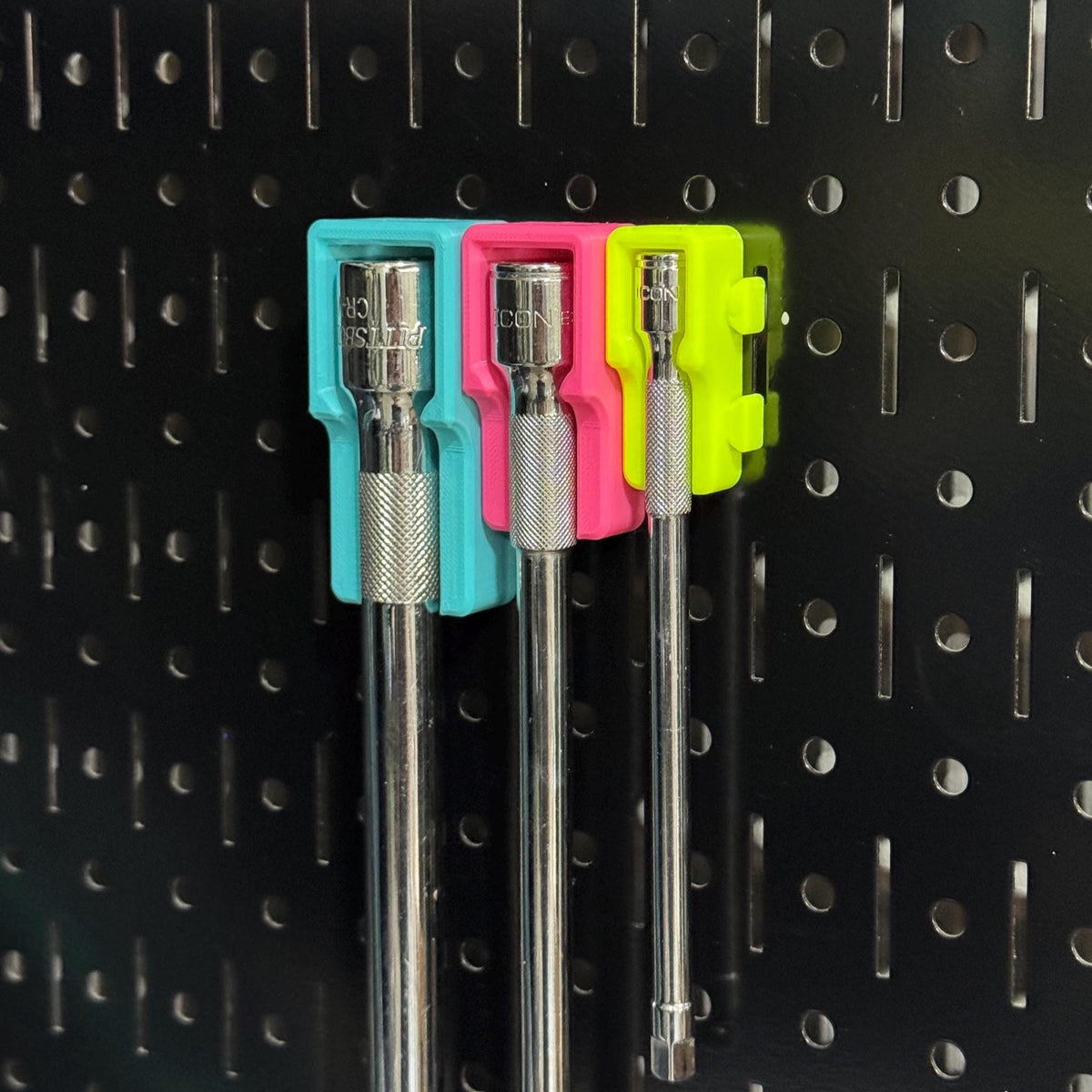 Three extension bars hanging side by side from teal, pink and yellow holders on black pegboard.
